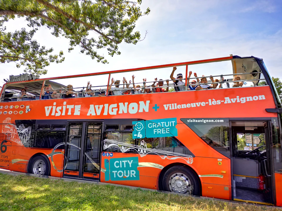 A bright red double-decker tour bus with people on the top deck, labeled "Visite Avignon + Villeneuve-lès-Avignon."