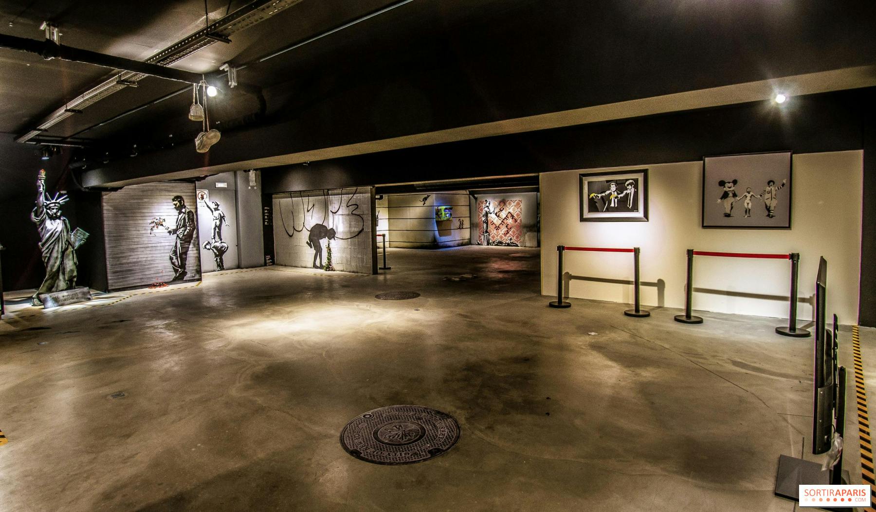 Una galleria poco illuminata che espone arte di strada, tra cui murales, stampe incorniciate e una statua, con barriere che delimitano alcune aree.