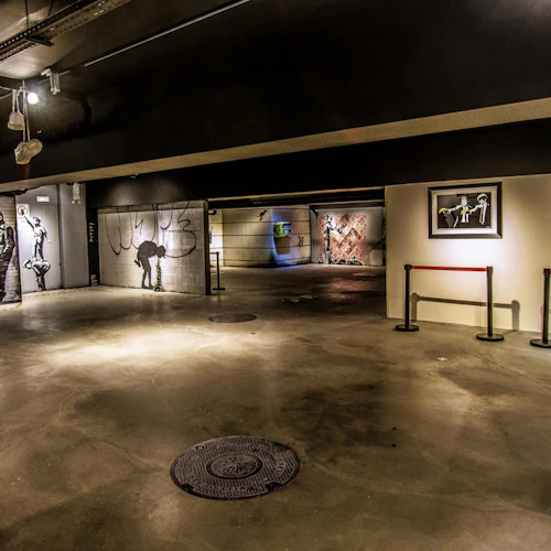 Ett svagt upplyst galleri som visar gatukonst, inklusive väggmålningar, inramade tryck och en staty, med barriärer som spärrar av vissa områden.