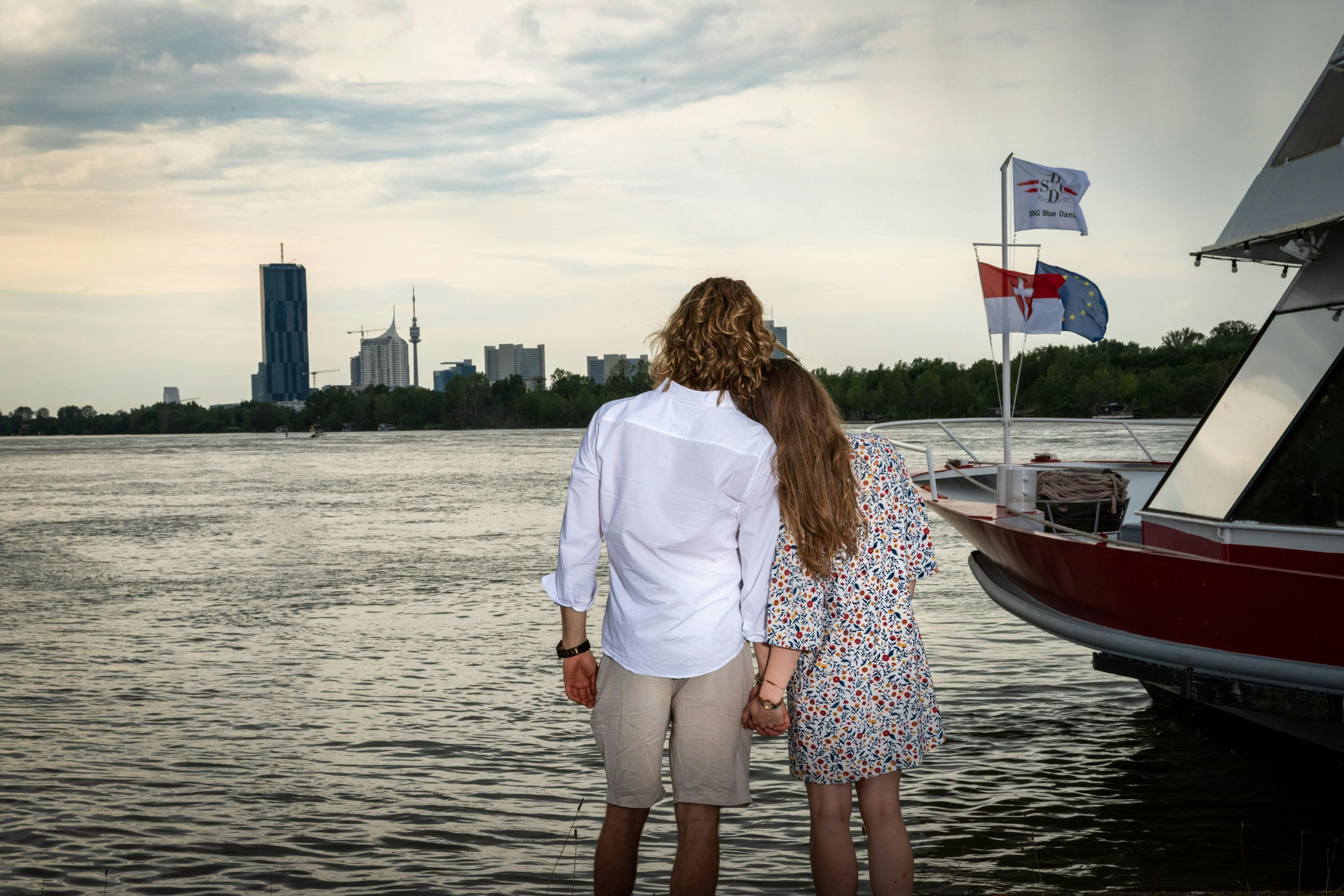 A couple stands by a river holding hands, facing city buildings in the distance. A boat with flags is on the water.