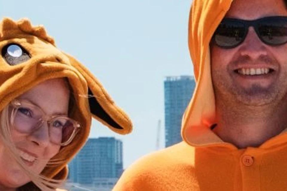 Two people wearing animal-themed onesies pose for a photo on a sunny day near a waterfront and a city skyline.