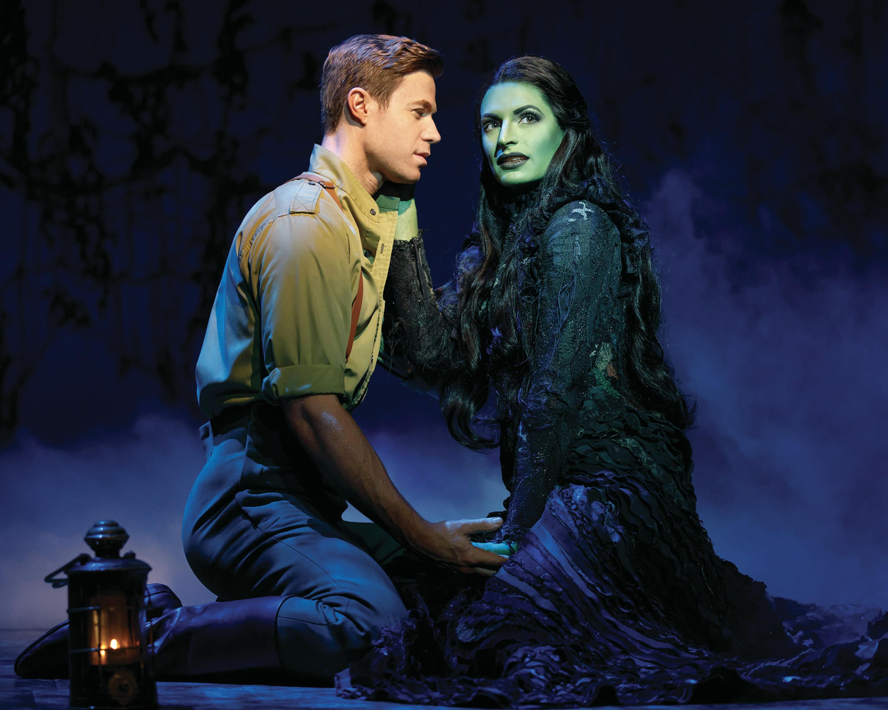 一名身穿棕褐色军装的男子站在黑暗、朦胧的舞台上，手持一张绿色皮肤、黑色长发女士的脸。