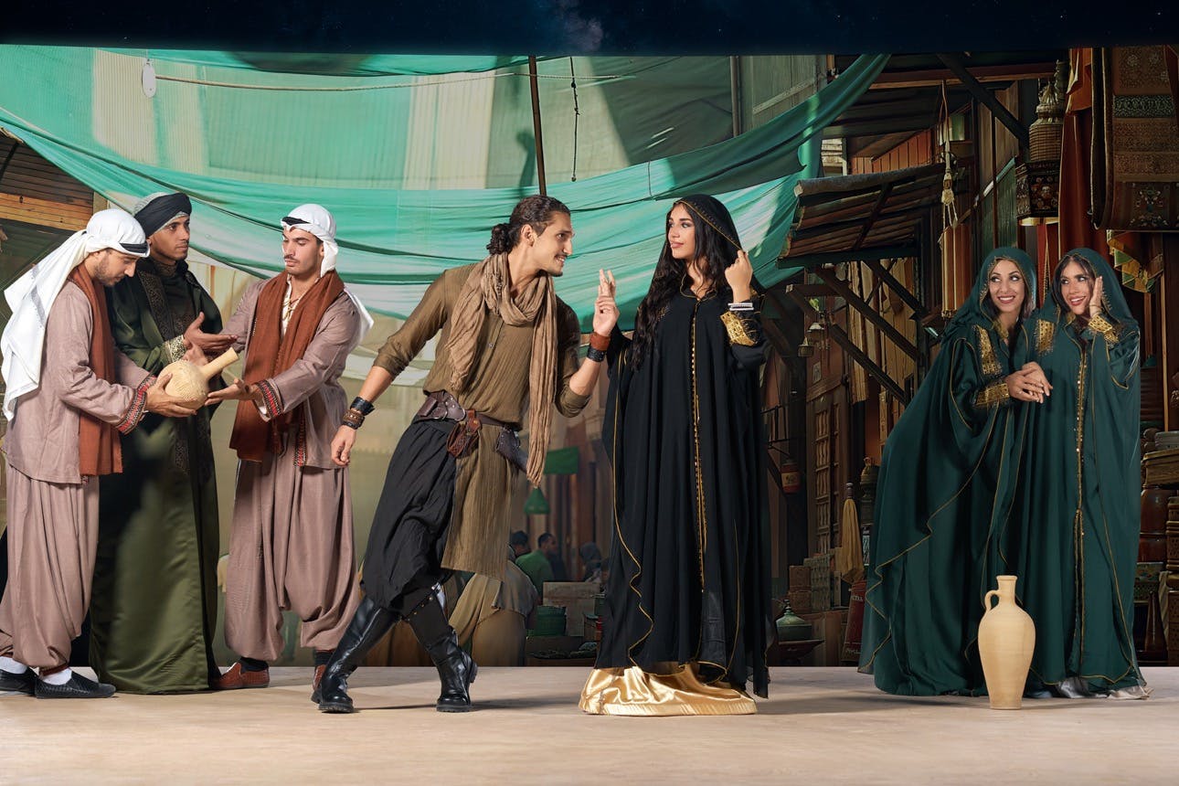 Five individuals in traditional Middle Eastern attire, three men on the left passing a jar, and a man and woman on the right gesturing.