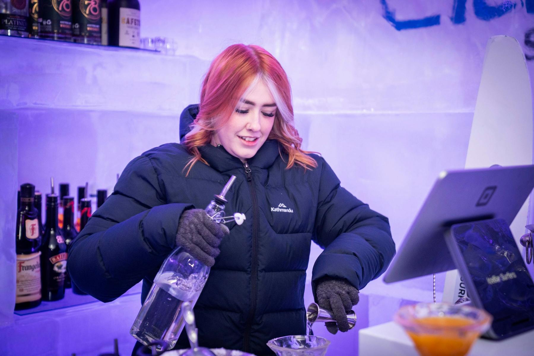 주황색과 흰색 머리카락을 가진 무거운 재킷을 입은 사람이 얼음 벽이 있는 바의 병에서 음료를 쏟아냅니다.