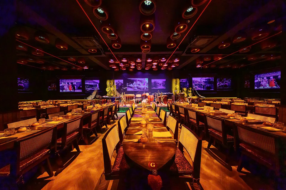 Dimly lit restaurant with long tables set for dining, illuminated by warm lighting. Purple-lit stage with musical instruments in the background.