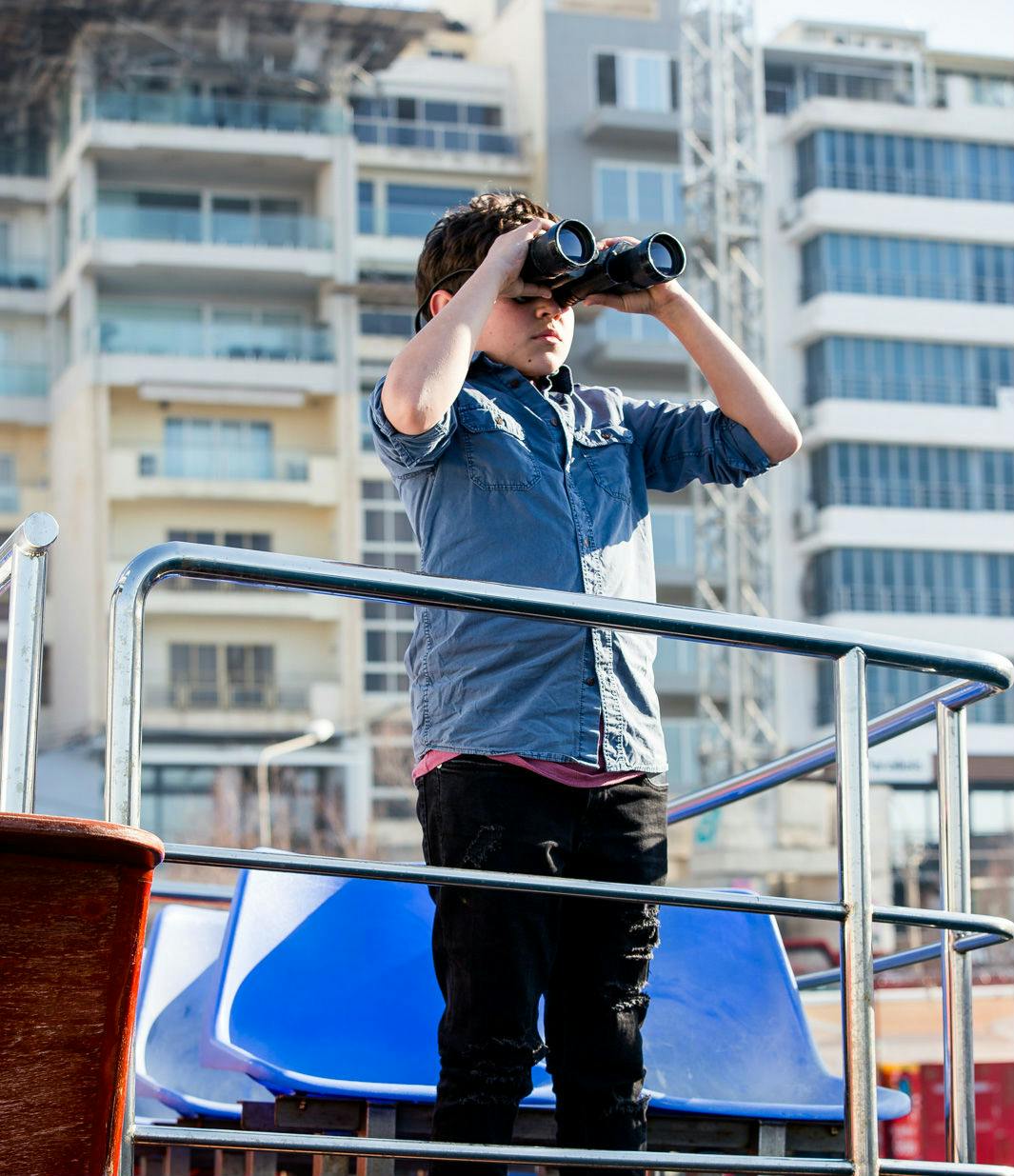 ボートのデッキに立っている少年が、建物を背景にした双眼鏡を通して眺めています。