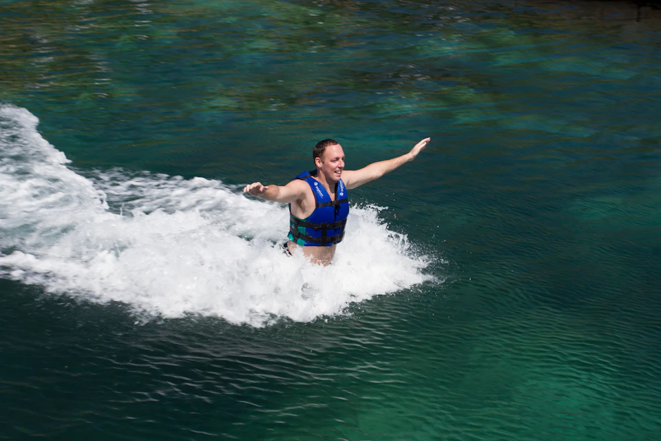 A man wearing a blue life vest is being propelled forward in a body of water, creating splashes around him.
