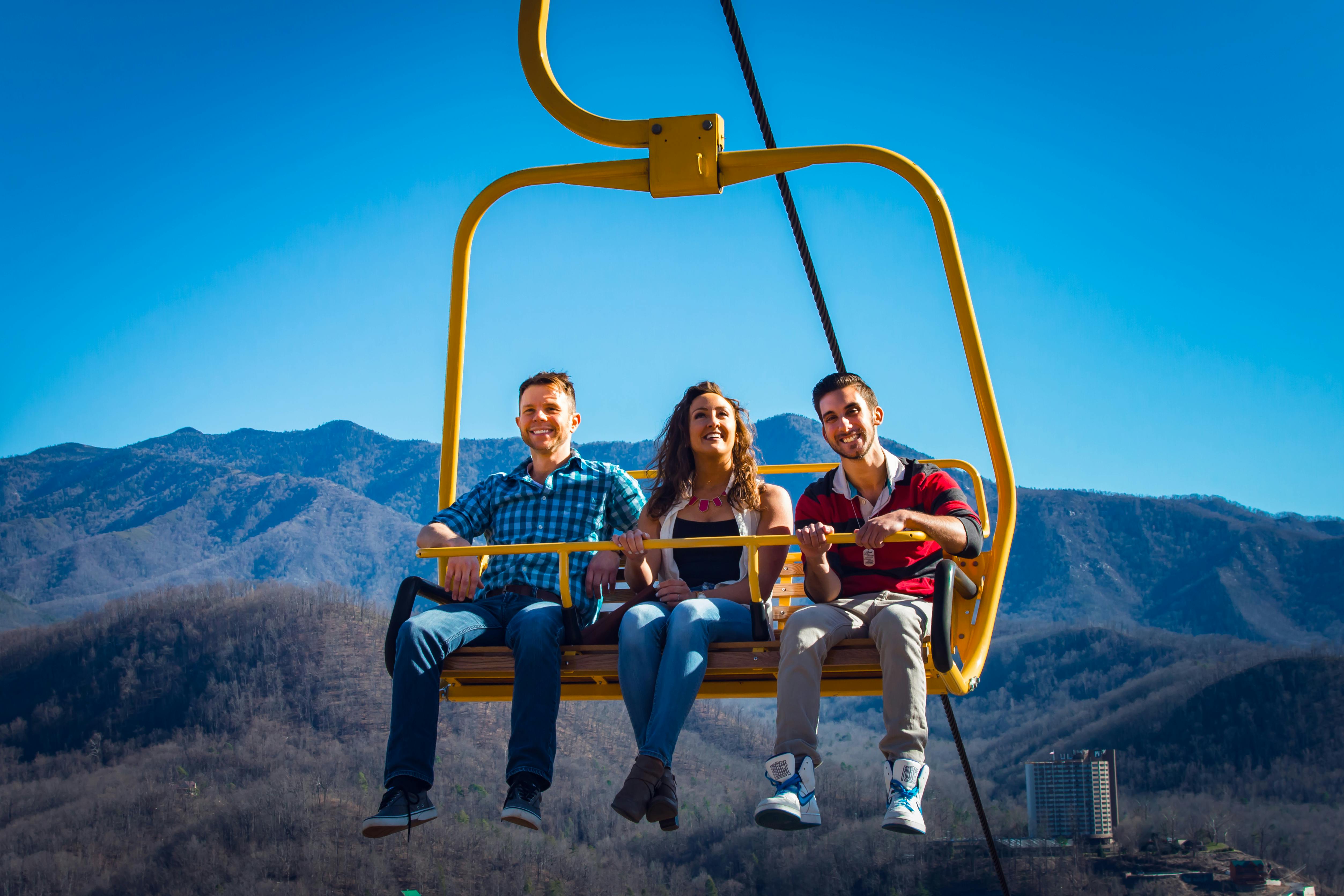 Tři lidé se usmívají a jezdí žlutým lyžařským vlekem s horskou scenérií v pozadí pod jasnou modrou oblohou.