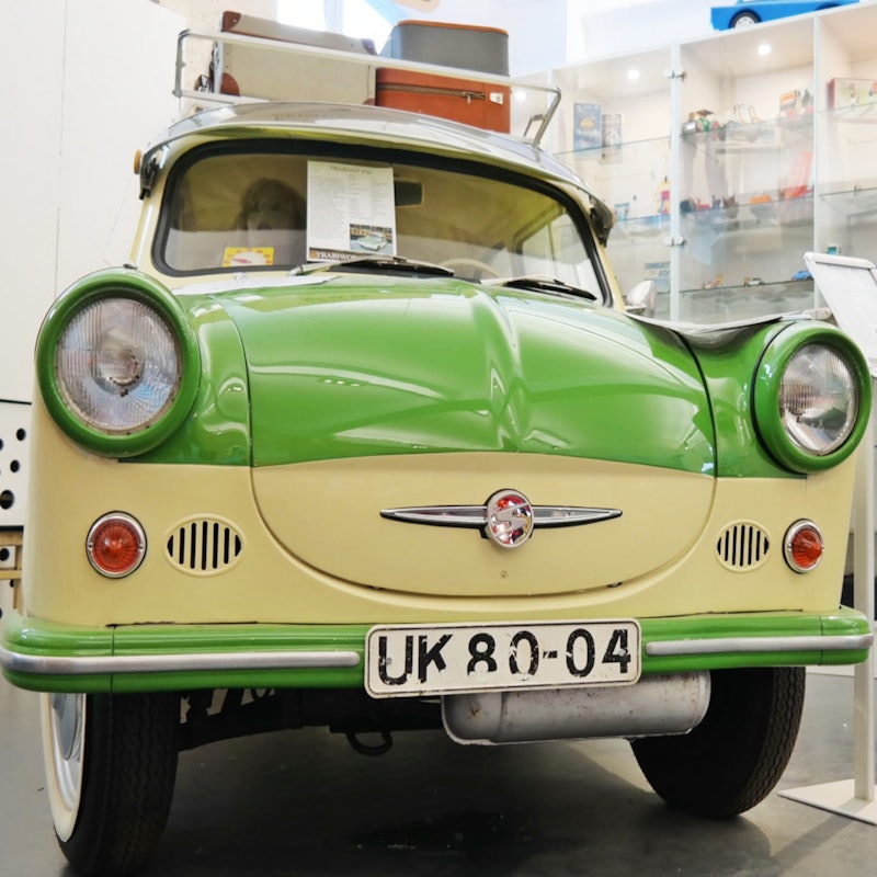 Vintagegrøn og cremefarvet bil udstillet på et museum med nummerplade "UK 80-04" og forskellige artefakter i baggrunden.