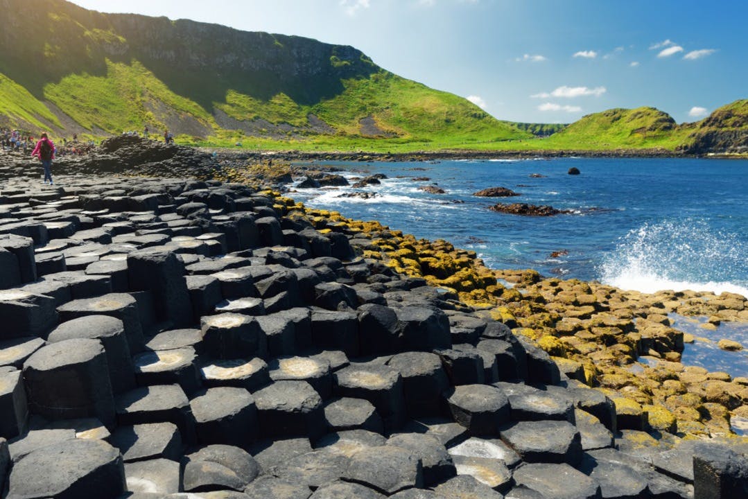 Colonnes hexagonales de basalte menant à la mer, avec des collines vertes et un ciel bleu en arrière-plan.