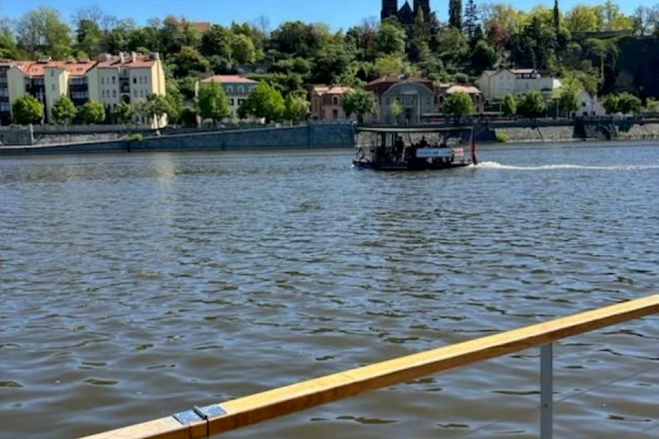 A river with a ferryboat, surrounded by buildings and greenery, under a clear blue sky.
