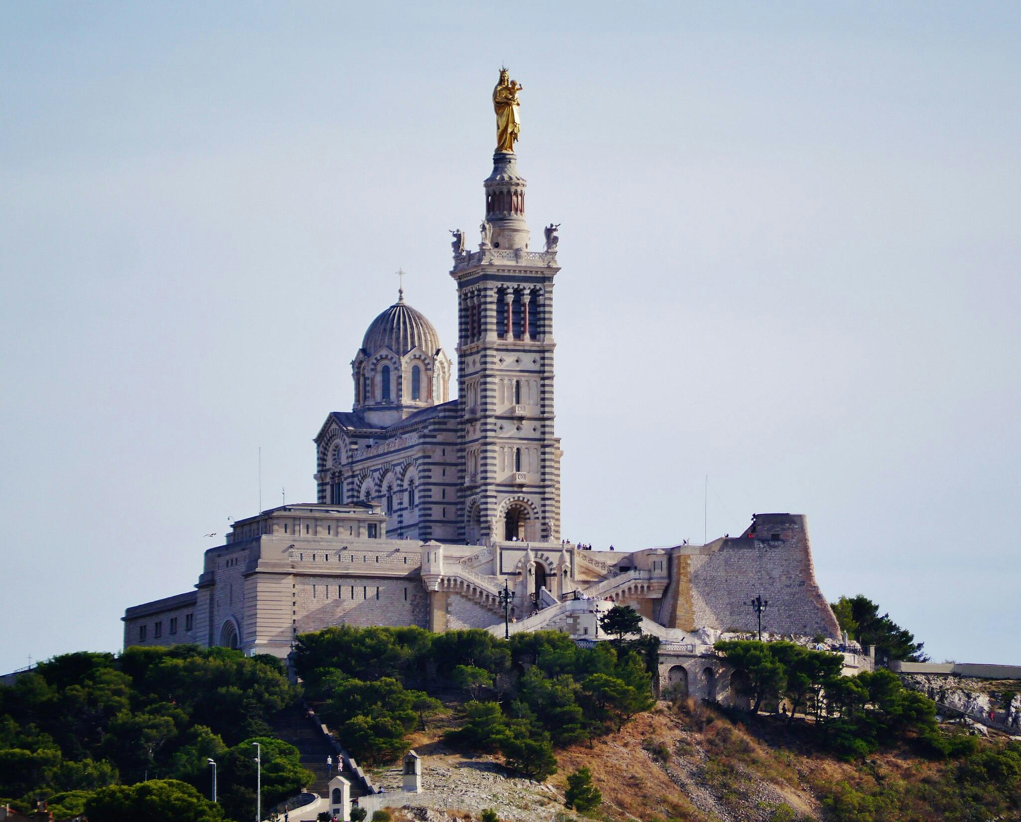 Na kopci obklopeném zelení pod jasnou oblohou stojí ozdobná bazilika s vysokou věží a zlatou sochou.
