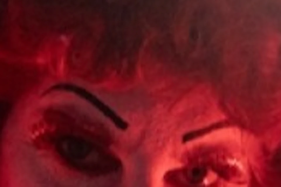 Person dressed as a character with red curly hair, a crown made of playing cards, red costume, and intense makeup under red lighting.