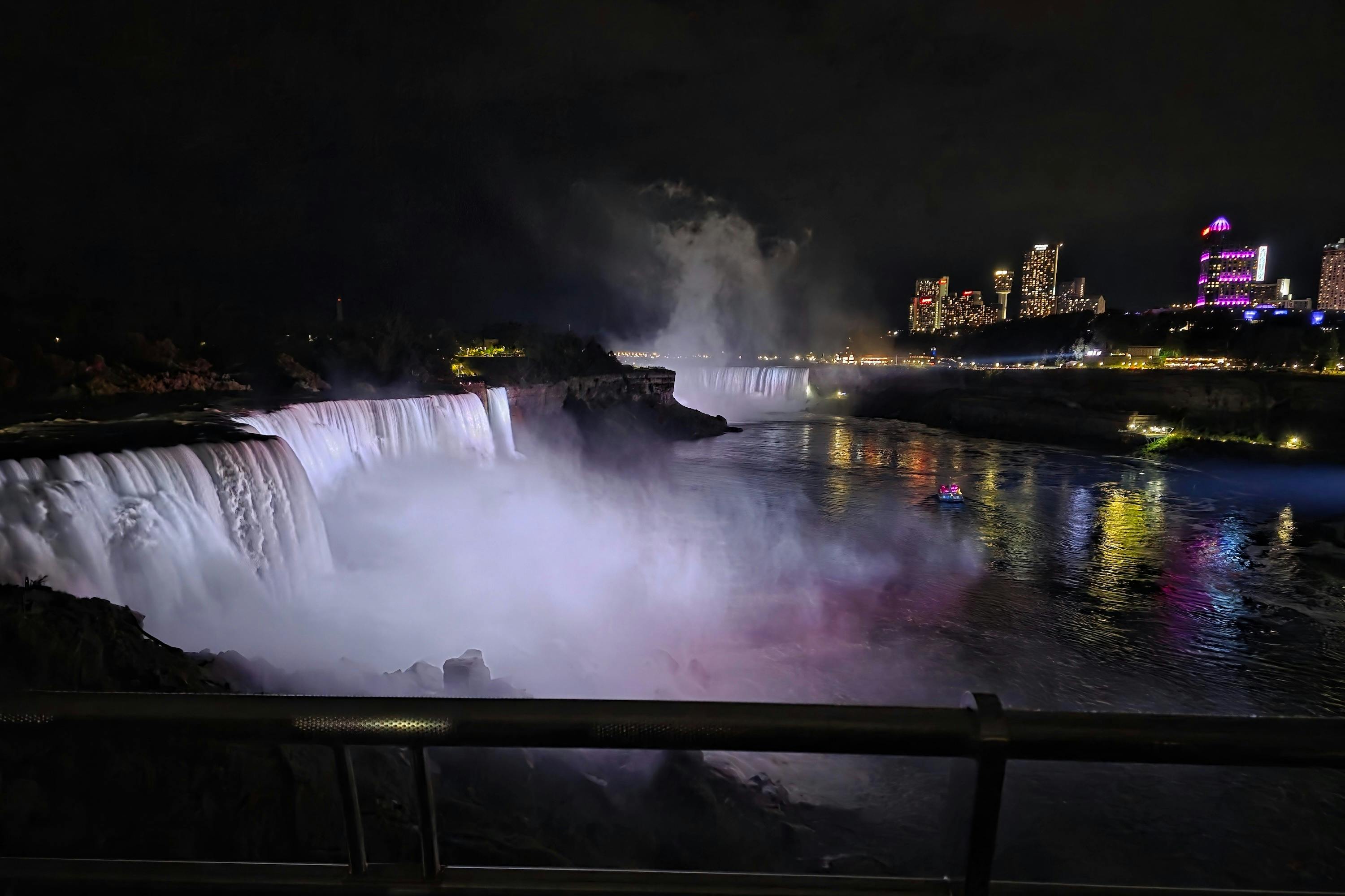 夜景中可以看到灯火通明的瀑布和升起的薄雾，背景是城市天际线，水下还有一艘船。