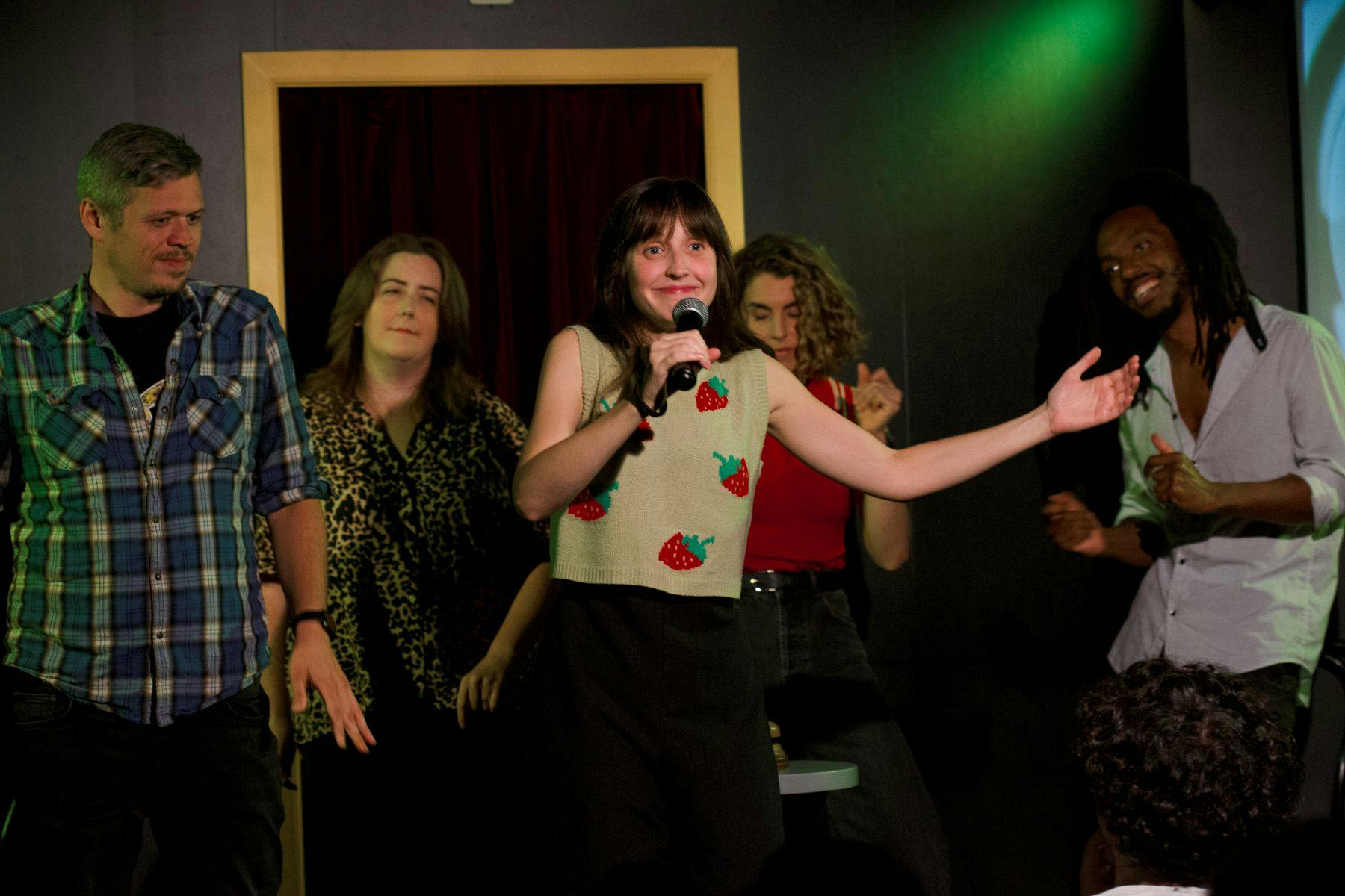 Eine Gruppe von vier Personen auf einer Bühne. Eine Person im Vordergrund hält ein Mikrofon und spricht oder spielt.