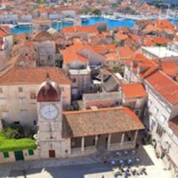 Flygfoto över en stad med många byggnader med röda tak och ett klocktorn, med en småbåtshamn och båtar i bakgrunden.