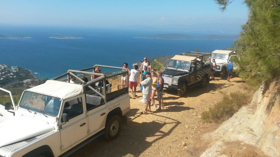 A group of people stands near safari jeeps on a dirt road overlooking a vast body of water and distant islands.