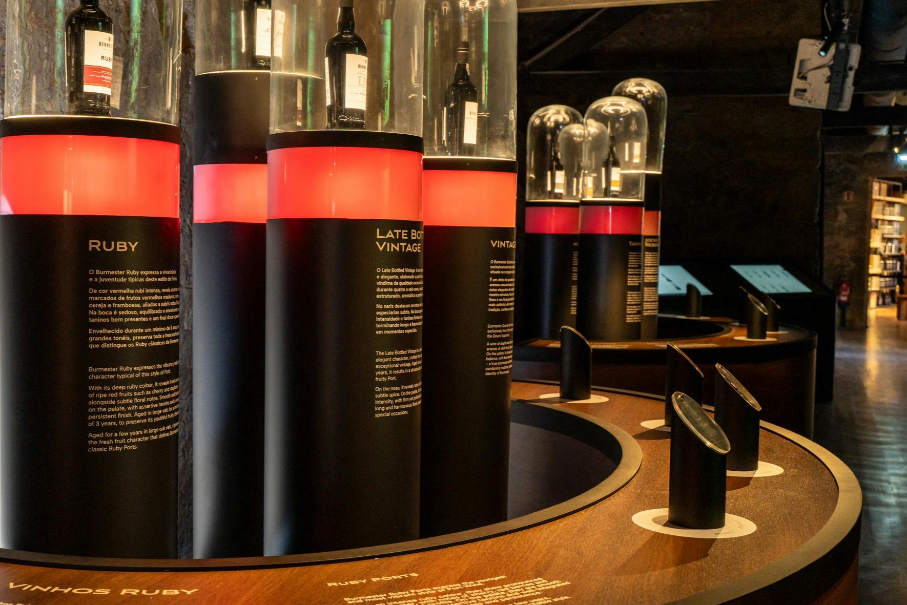 Exposition de bouteilles présentées sous des dômes de verre sur des colonnes lumineuses avec des descriptions.