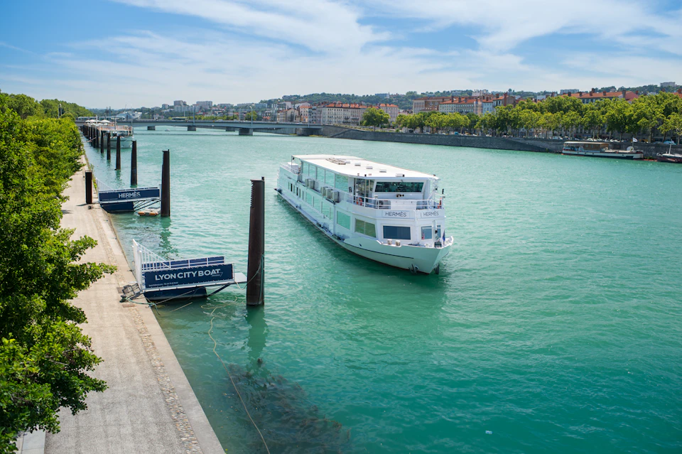 A white boat labeled "HERMES" floats on a green river near a dock labeled "LYON CITY BOAT" under a blue sky.