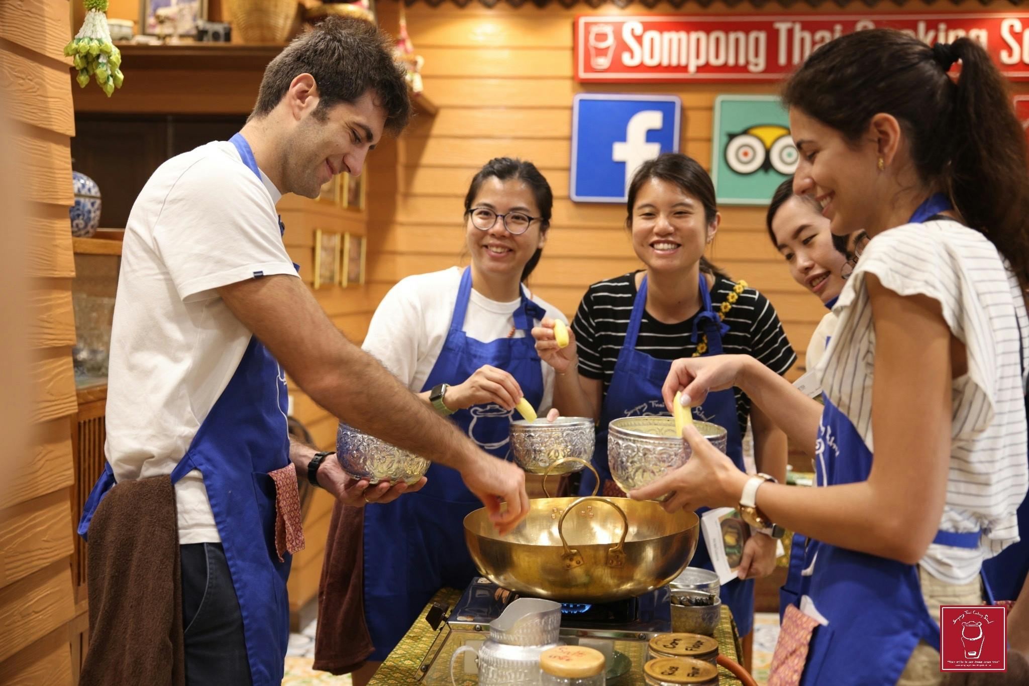A group of people in blue aprons cooking together, holding utensils and smiling in a cozy kitchen setting.