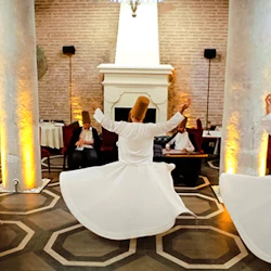 Tre män i vita kläder och koniska hattar som virvlar runt i en dans, med musiker som spelar sittande i bakgrunden.