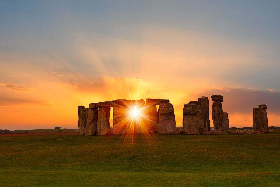 Stonehenge at sunset with the sun perfectly aligned between two stones, casting vibrant orange and yellow hues.