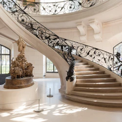 Elegant spiraltrappa med utsmyckade svarta räcken i ett ljust rum, med en stor, invecklad staty vid basen.