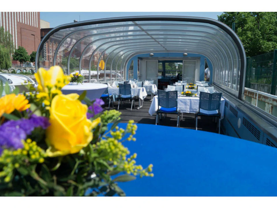 A boat with a transparent roof has tables set with white cloths and blue chairs, adorned with yellow flowers.
