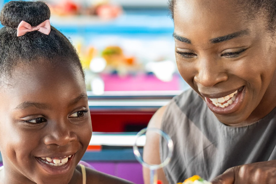 A woman and a child smiling while seated at a table with colorful food platters and desserts in a brightly lit diner.