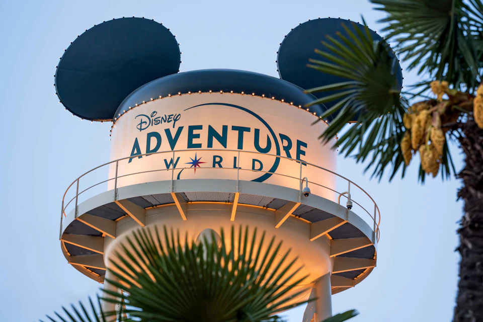 A water tower bearing the words "Disney Adventure World" and Mickey Mouse ears on top, surrounded by palm fronds.
