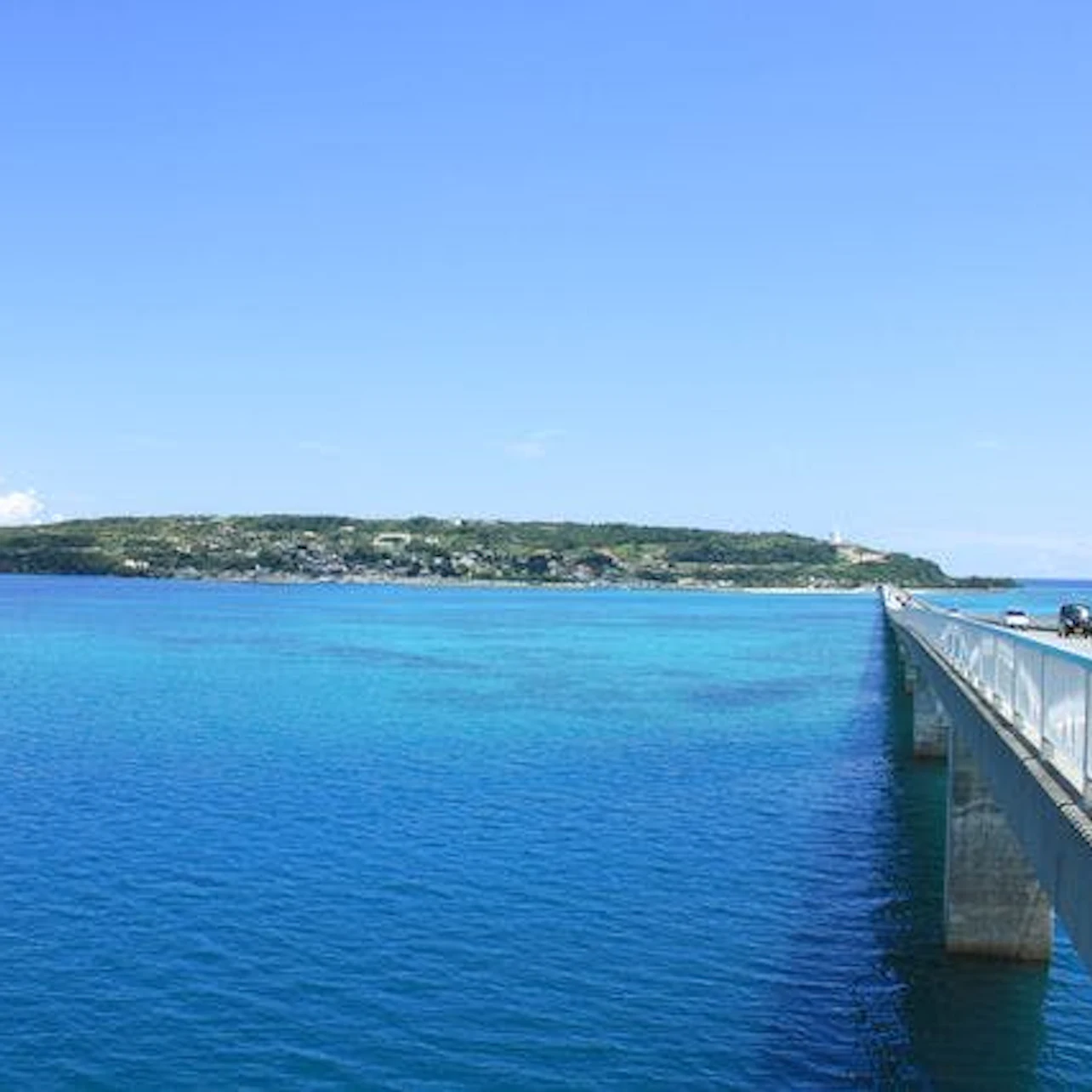A long bridge extends over clear, blue ocean water toward a distant, green landmass under a bright blue sky.