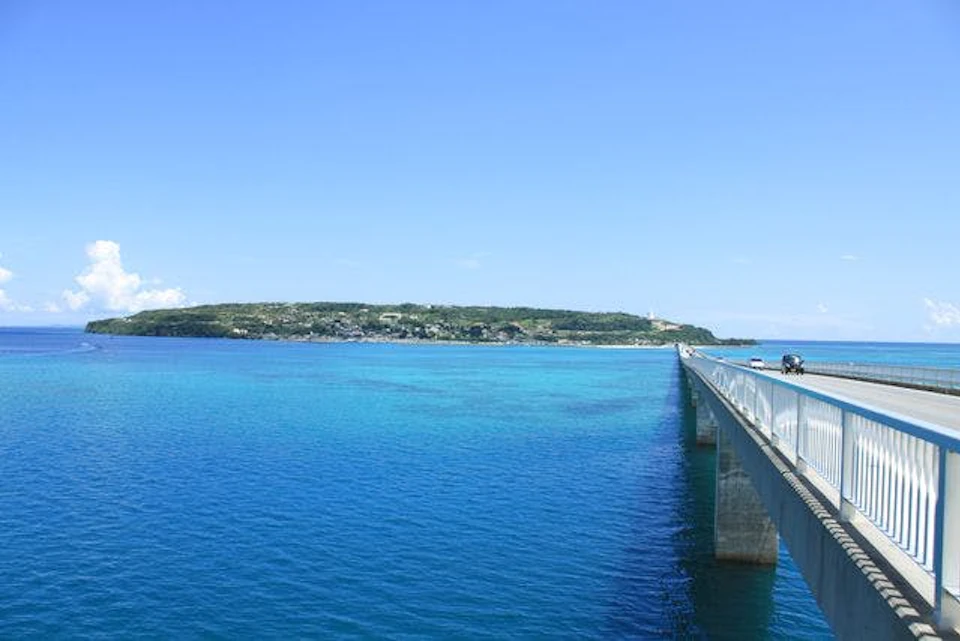 A long bridge extends over clear, blue ocean water toward a distant, green landmass under a bright blue sky.