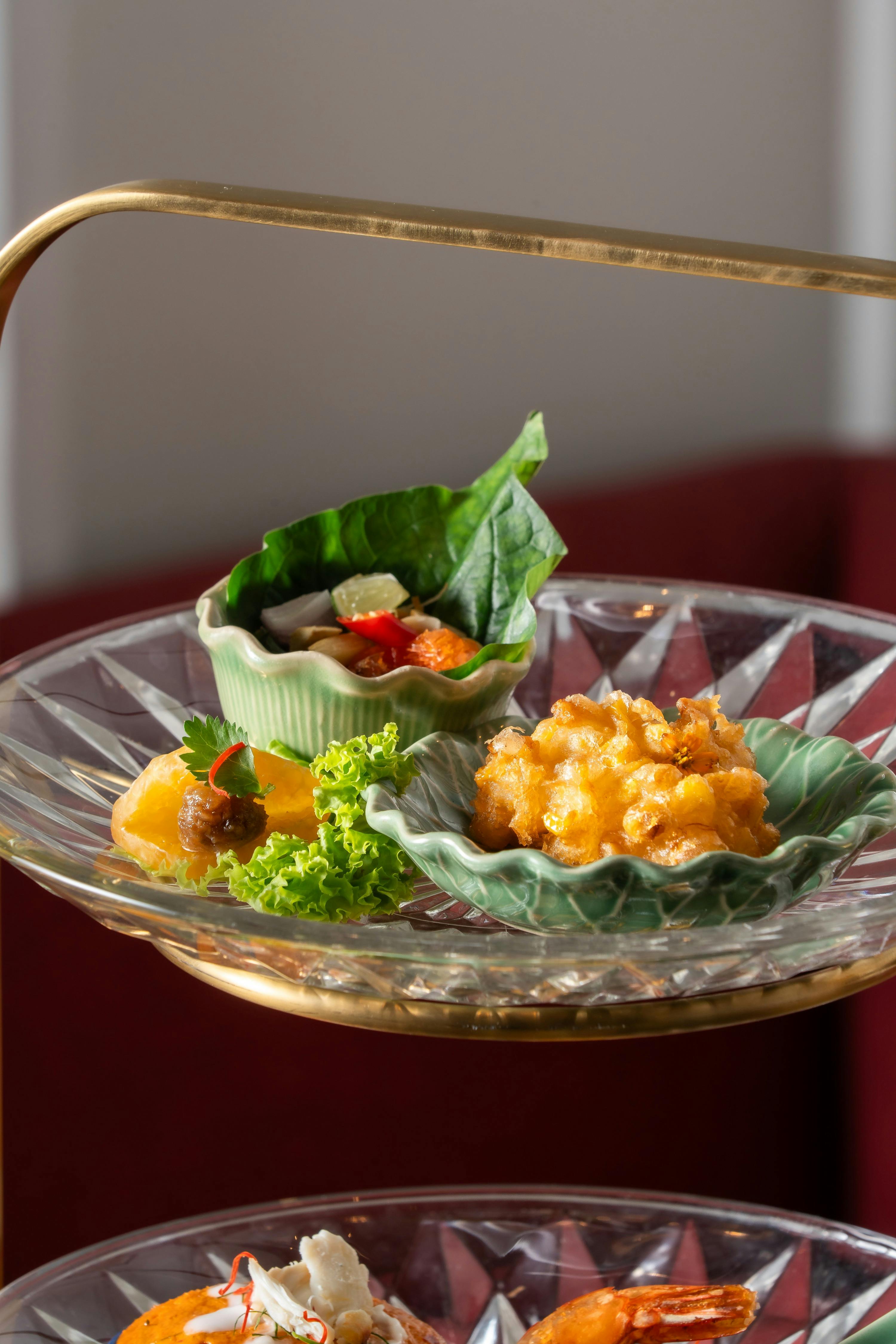 A glass plate with green ceramic bowls containing various small portions of food, including greens, vegetables, and fried items.