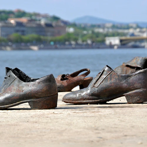 Flera par rostiga, gammaldags skor placeras på en betongvall bredvid en flod, byggnader och en bro i bakgrunden.