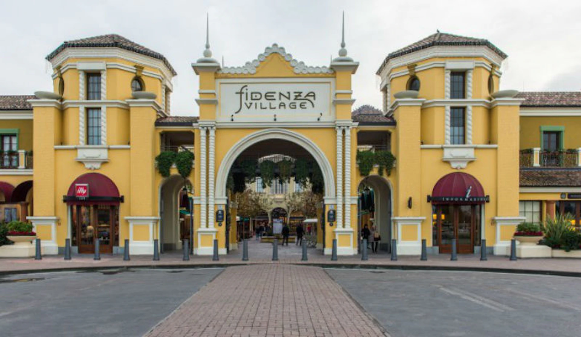 Yellow archway entrance labeled "Fidenza Village" with shops on either side and a cobblestone walkway.