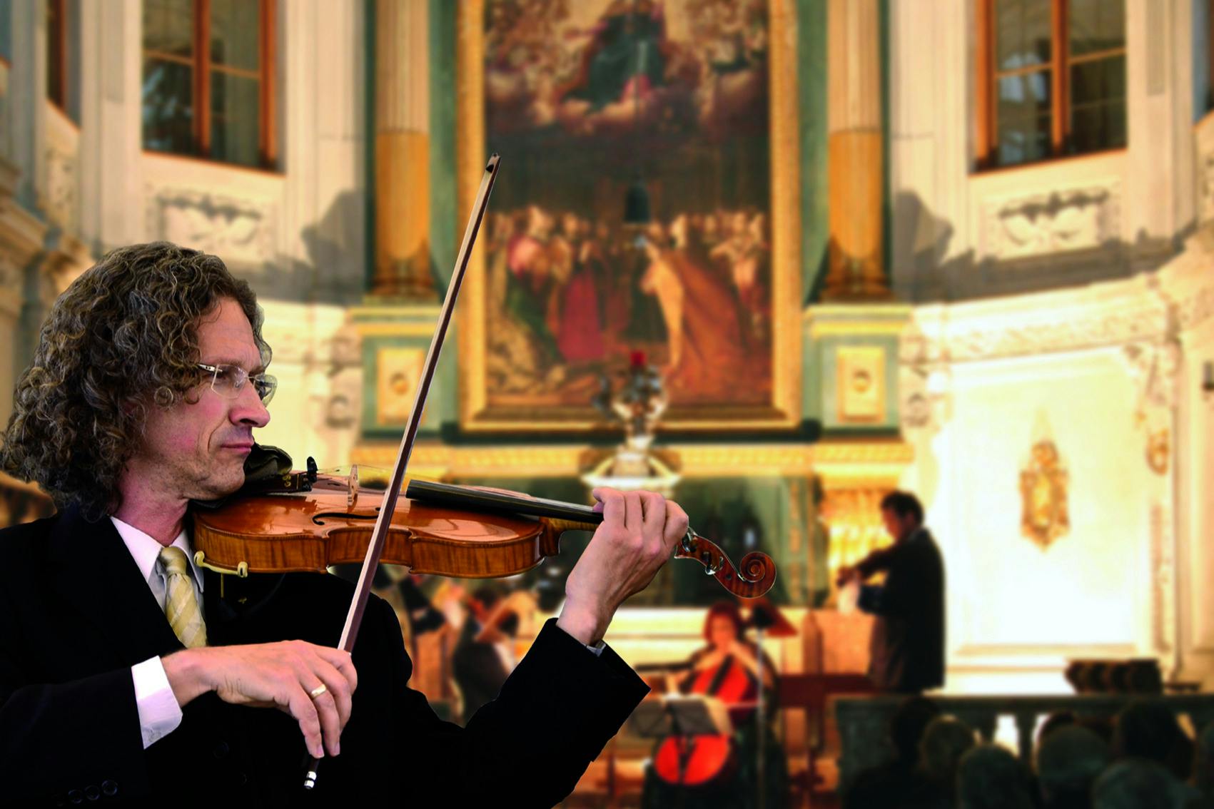 A man with curly hair and glasses plays a violin in an ornate, indoor setting. An audience and musicians are visible in the background.