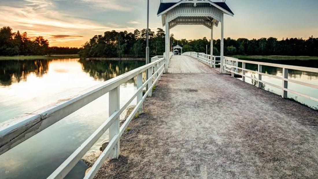 Un puente de madera con barandillas blancas y una parte cubierta se extiende sobre aguas tranquilas hacia una zona boscosa al atardecer.