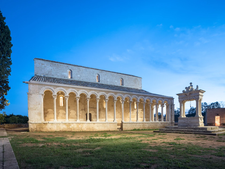 An ancient stone building with arched columns and lit interior, under a clear blue sky, with a decorative structure on the right.