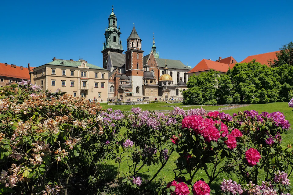 A historic cathedral with towers and domes, surrounded by greenery and blooming flowers under a clear blue sky.