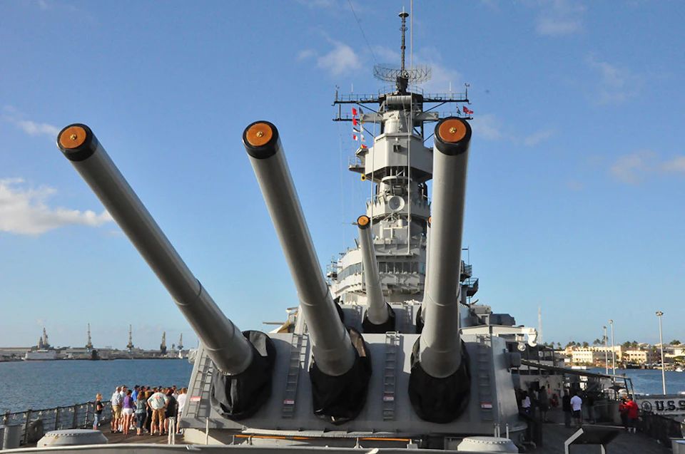 Large naval battleship with prominent gun barrels, docked at port. A group of people is gathered on the deck near the ship's bow.