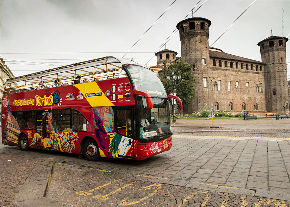 A red double-decker tour bus passes in front of an old stone building with towers and arches.