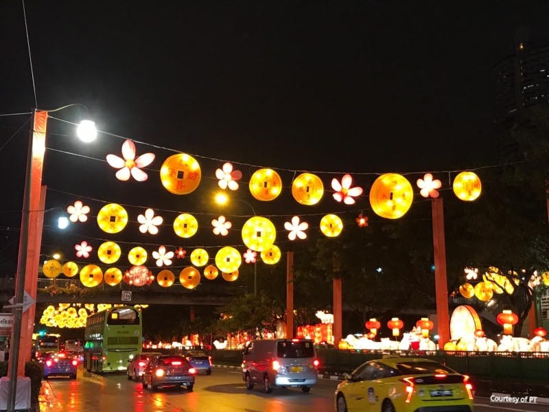 Ulice zdobená závěsnými lucernami a květinovými světly v noci, s vozidly včetně autobusu a taxi.