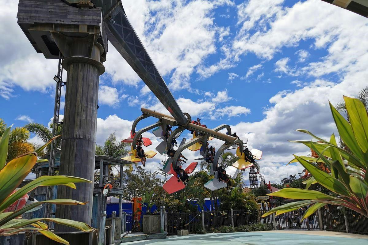 A suspended roller coaster with riders in colorful seats, set against a blue sky with clouds and surrounding greenery.