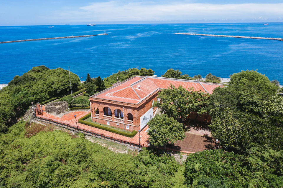 A red-roofed building surrounded by greenery near a coastline with a clear blue sea and a bright sky.