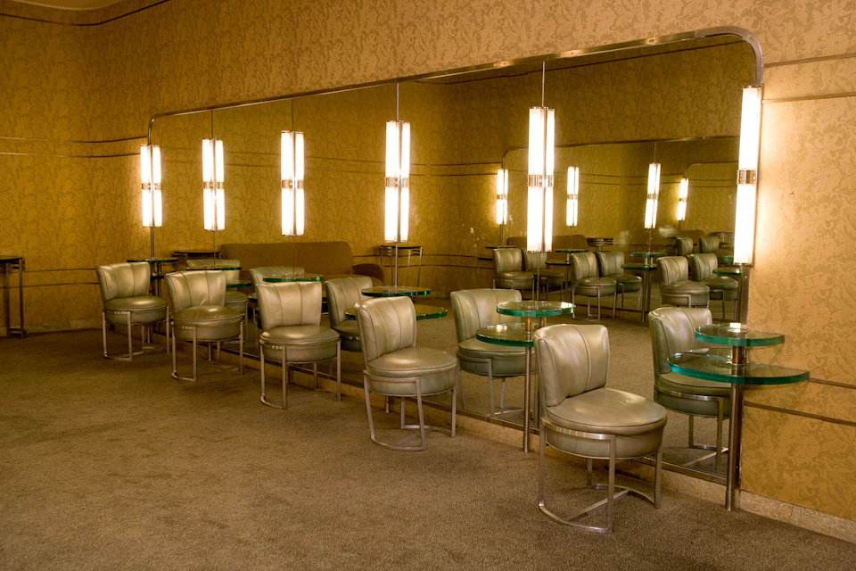 Retro-style lounge with green leather chairs, round glass tables, and illuminated wall lights reflecting in large mirrors.