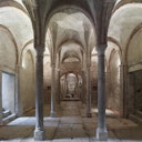 A historical stone crypt with arched ceilings and columns, featuring steps on the left and a decorative gate at the far end.