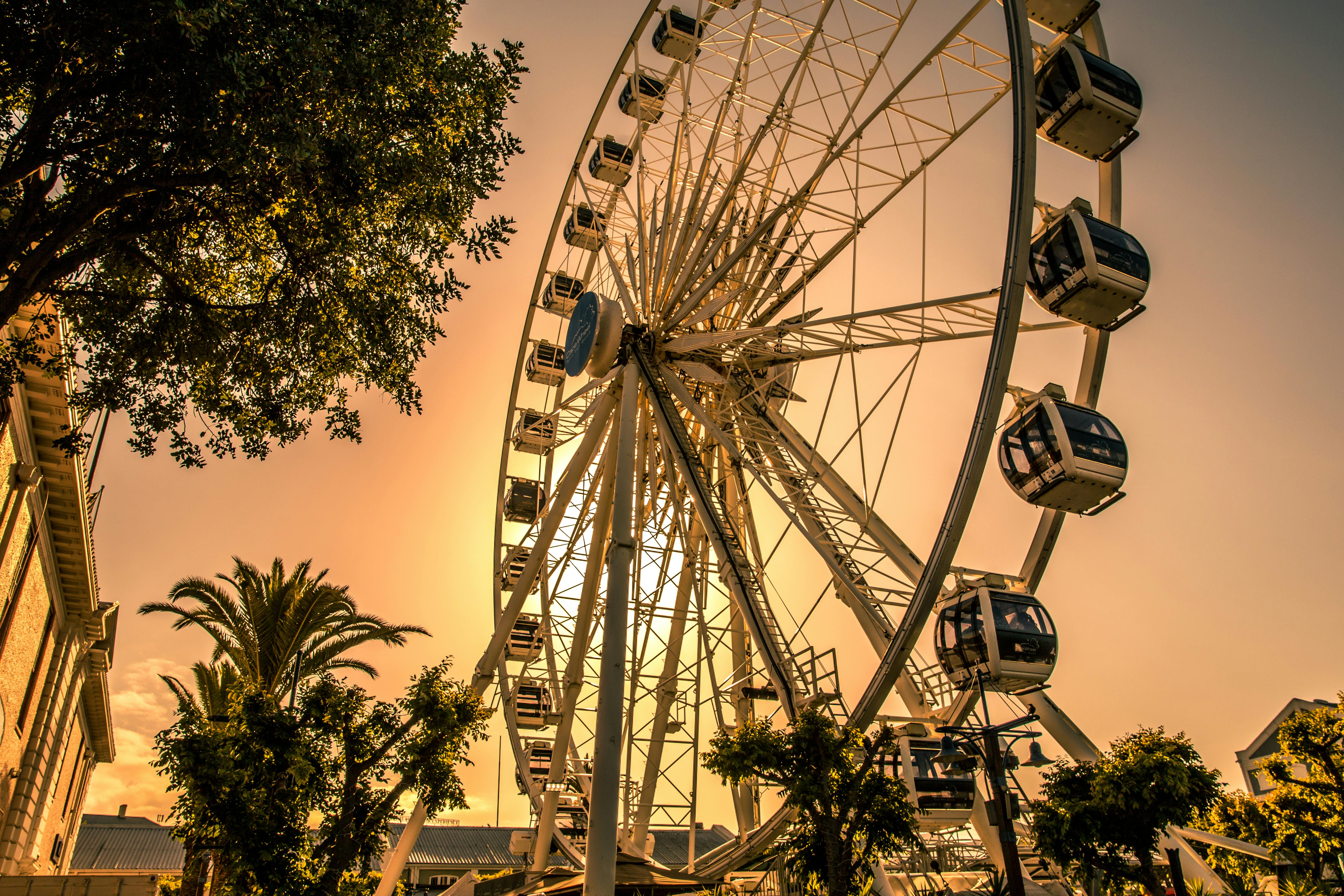 Une grande roue avec des cabines fermées sur fond de ciel doré, entourée d'arbres et d'un bâtiment sur la gauche.