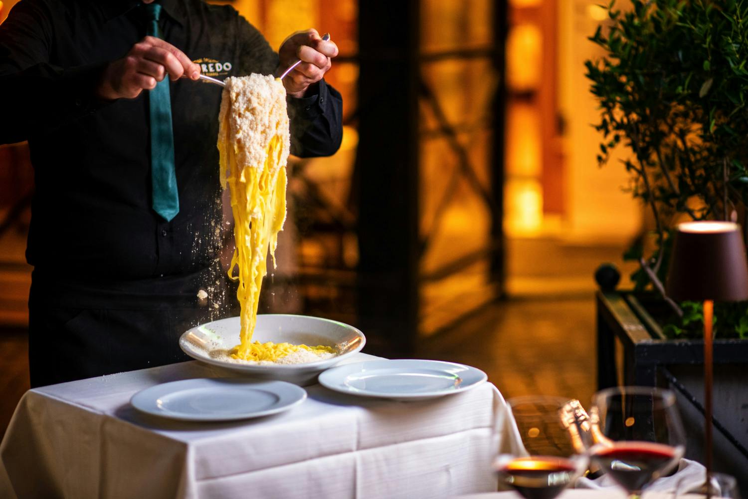 Человек в темной форме подает за столом пасту, покрытую сыром, с белыми тарелками и белой скатертью.