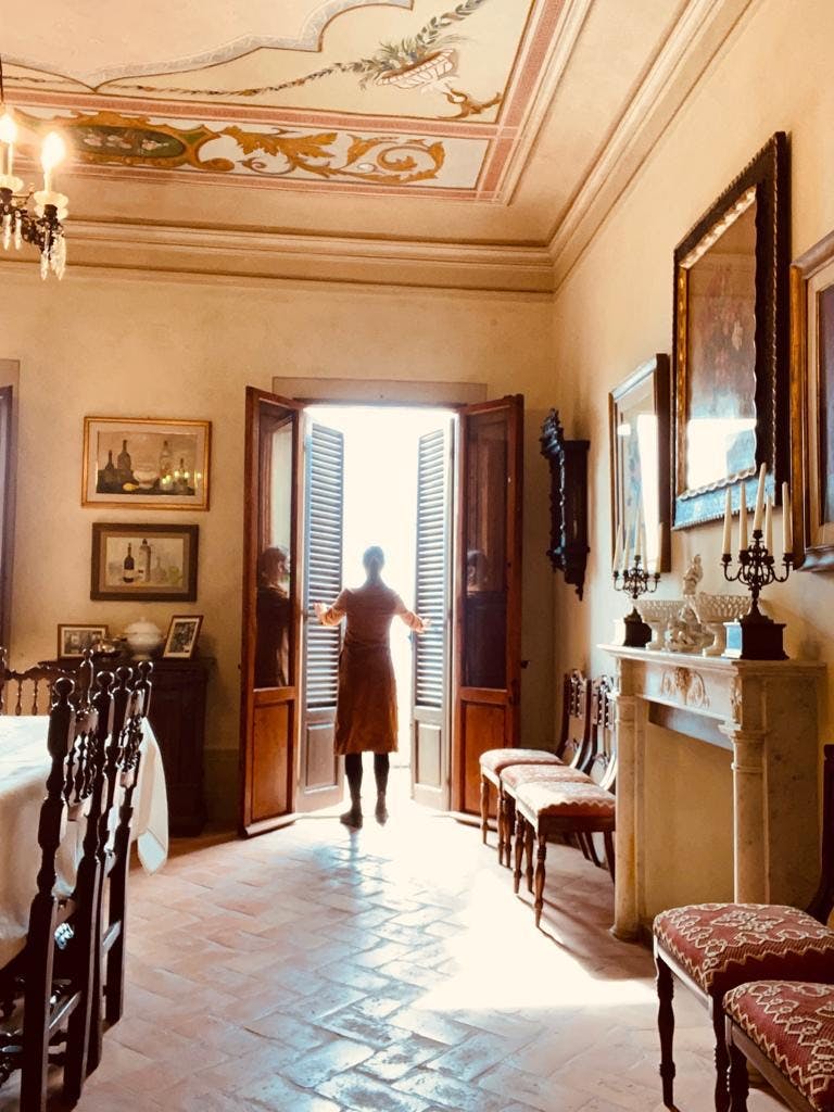 Una persona si trova in una stanza elegantemente decorata e apre le doppie porte alla luce del sole. La stanza presenta opere d'arte, sedie e un caminetto.