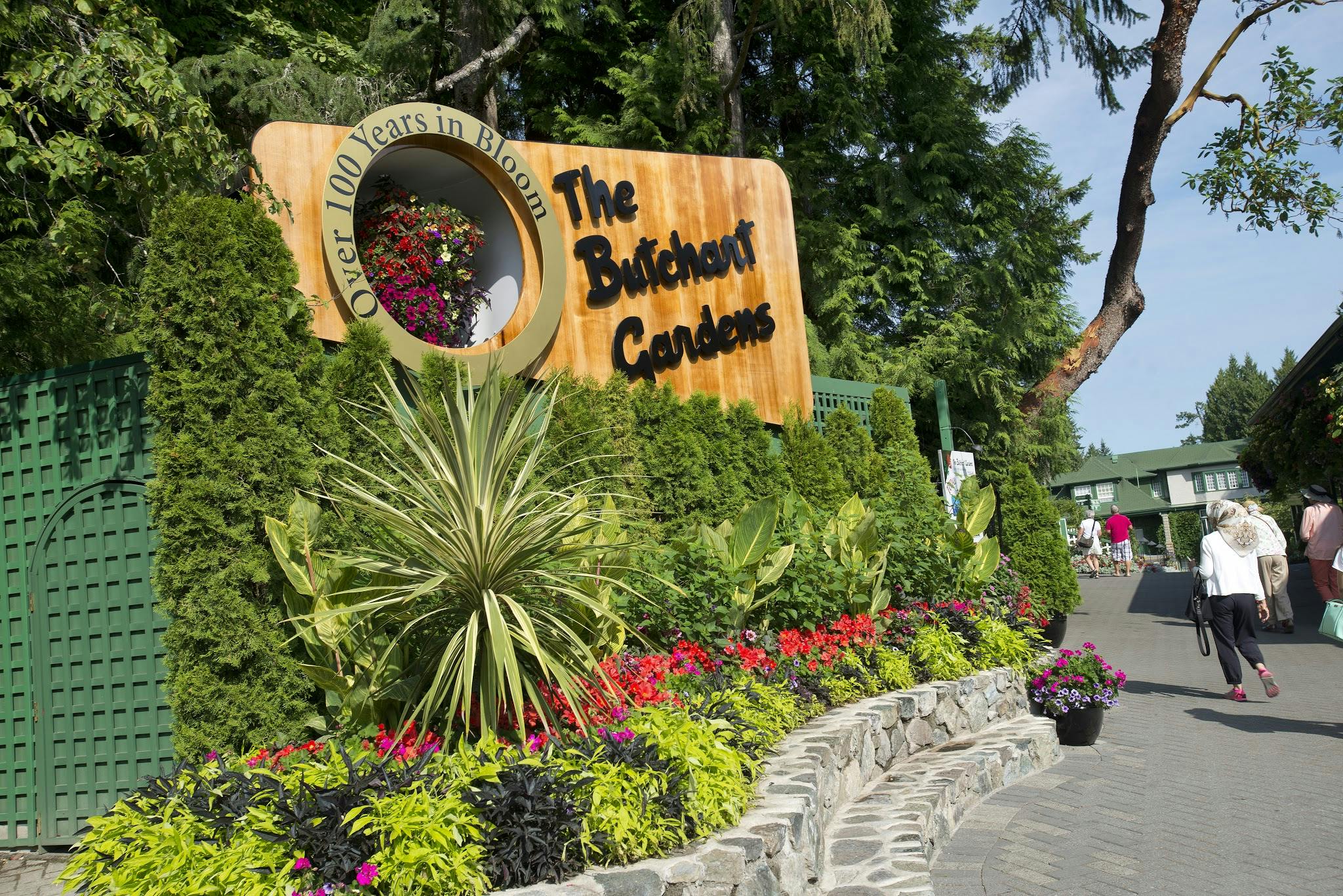 Πινακίδα εισόδου στους κήπους Butchart, που περιβάλλεται από καταπράσινο τοπίο και πολύχρωμα λουλούδια. Οι επισκέπτες φαίνονται στο παρασκήνιο.
